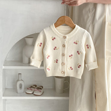 White children's cardigan with cherry embroidery on a wooden hanger against a neutral background.